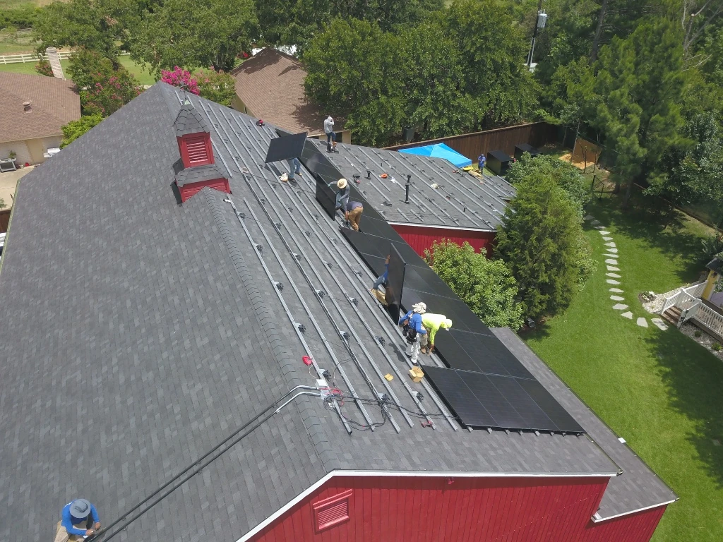 Solar panels installed on a gray shingled roof with a brick chimney in a residential neighborhood.