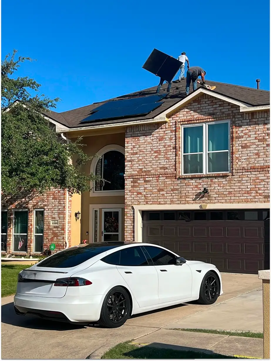 Two workers installing solar panels on the roof of a brick house with a white Tesla parked in the driveway.